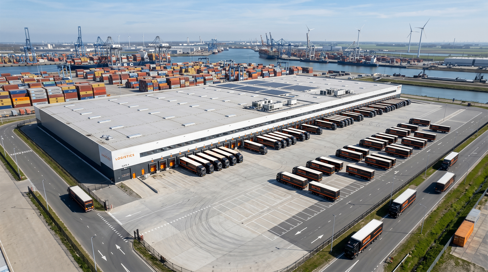 Aerial view of a large logistics warehouse beside Rotterdam port with containers, cranes, and trailers.